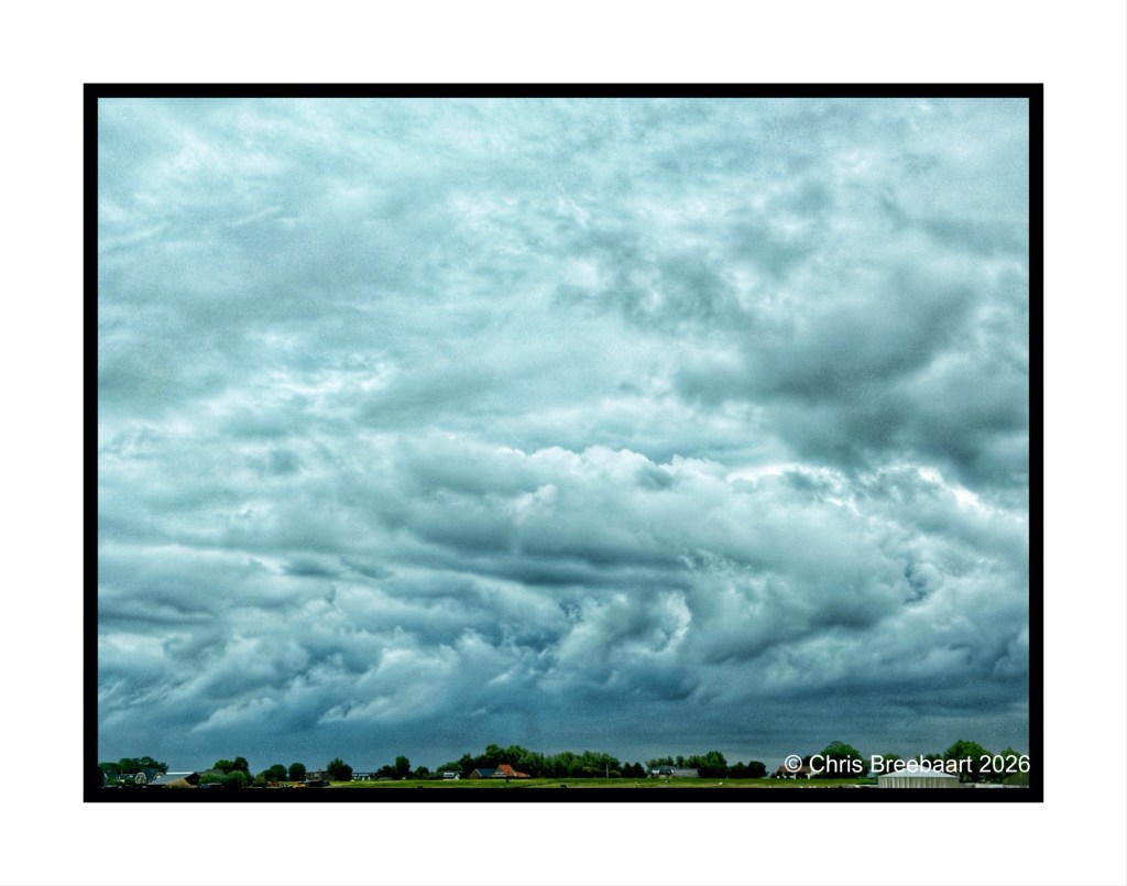 A dramatic sky filled with thick, gray clouds, with a hint of blue peeking through, over a rural landscape featuring fields and a few buildings in the foreground.