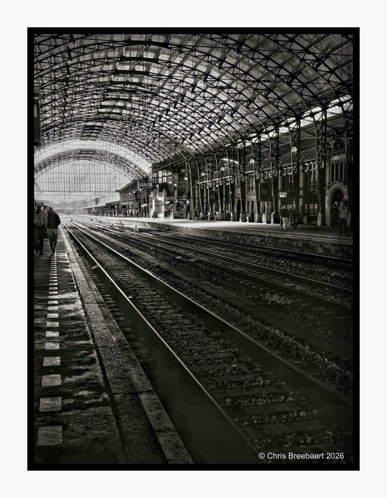 Black and white photograph of a railway station with a high arched roof, featuring train tracks leading into the distance and people walking along the platform.
