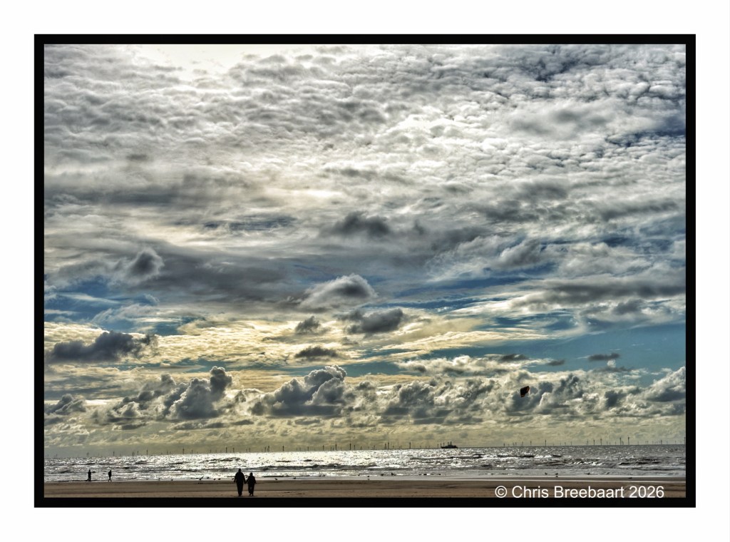 Couple walking along a beach under a dramatic sky filled with clouds, with the ocean in the background.