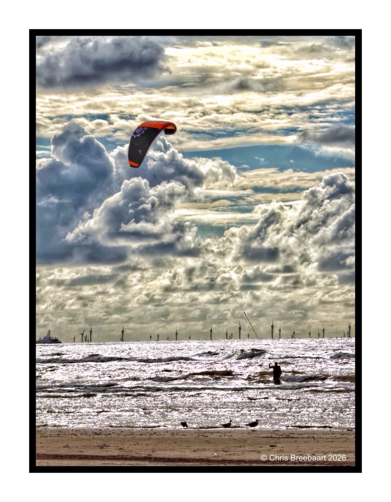 A kite surfer gliding through the air over shimmering ocean waves with dark clouds in the background and a silhouette of a fisherman standing in the water.