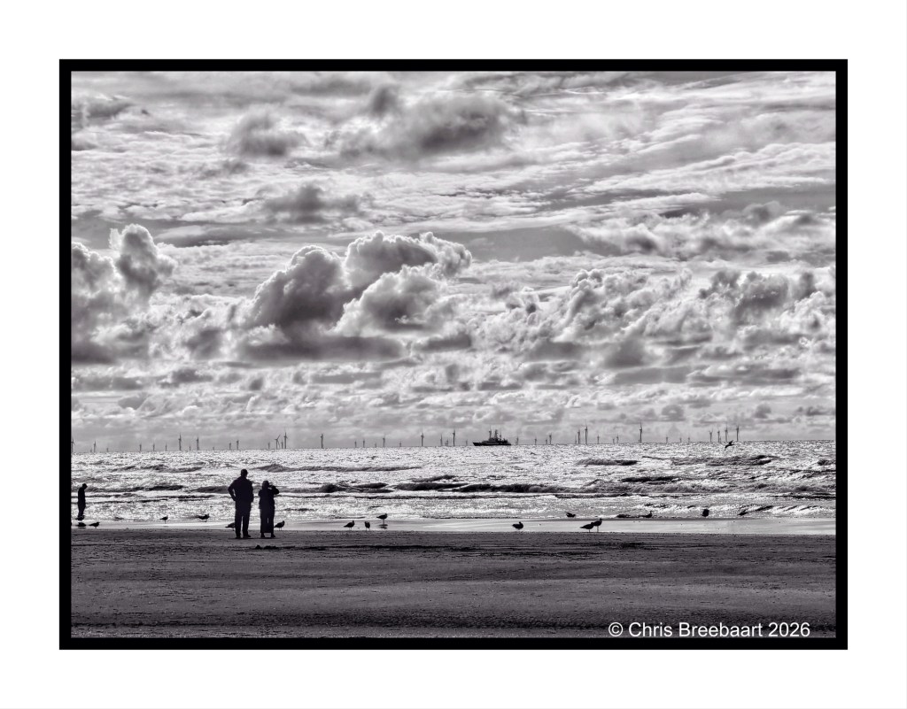 Black and white image of a beach with two people standing in the foreground, looking out toward the sea, where wind turbines are visible on the horizon under a cloudy sky.