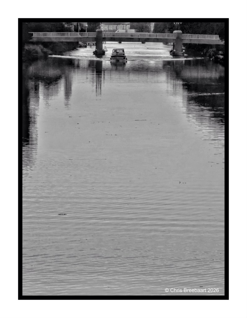 Black and white image of a calm river with a small boat navigating towards a bridge in the background.
