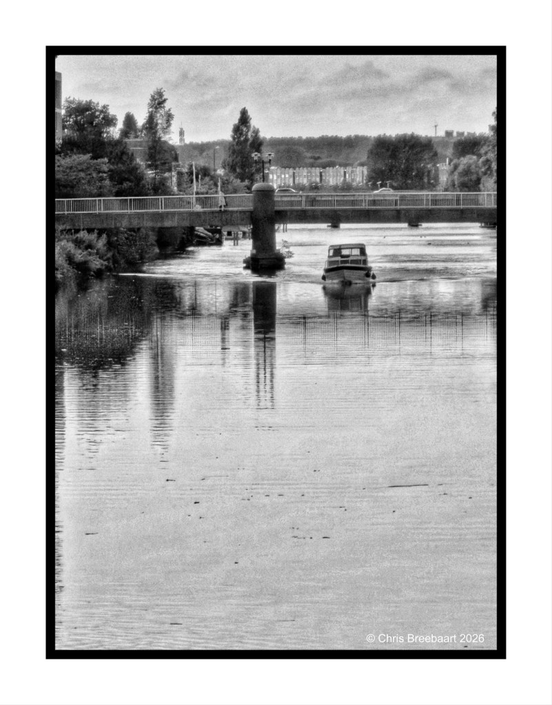 A black and white image of a river with a small boat navigating through it, under a bridge. The scene includes trees and buildings in the background, reflecting on the water's surface.