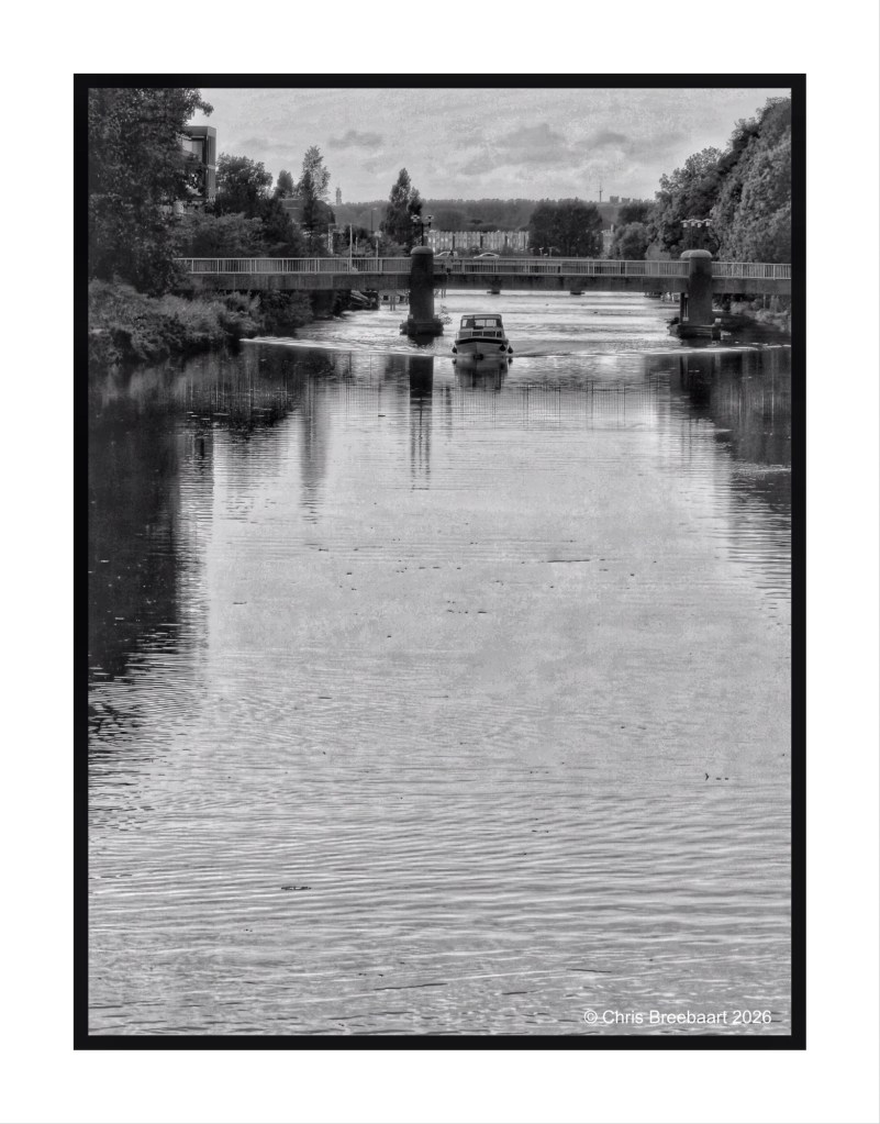 A black and white photograph of a river with a small boat navigating towards a bridge, surrounded by trees and buildings under a cloudy sky.