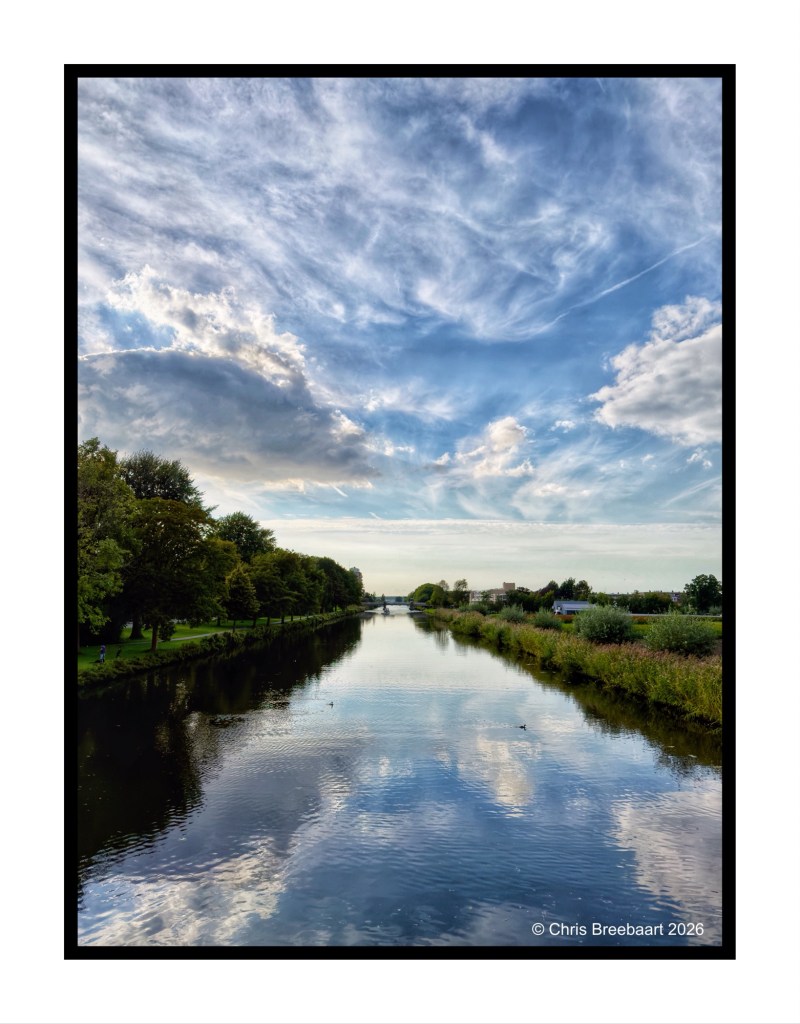A serene view of a river bordered by trees, reflecting the cloudy sky overhead with soft blue and white hues.