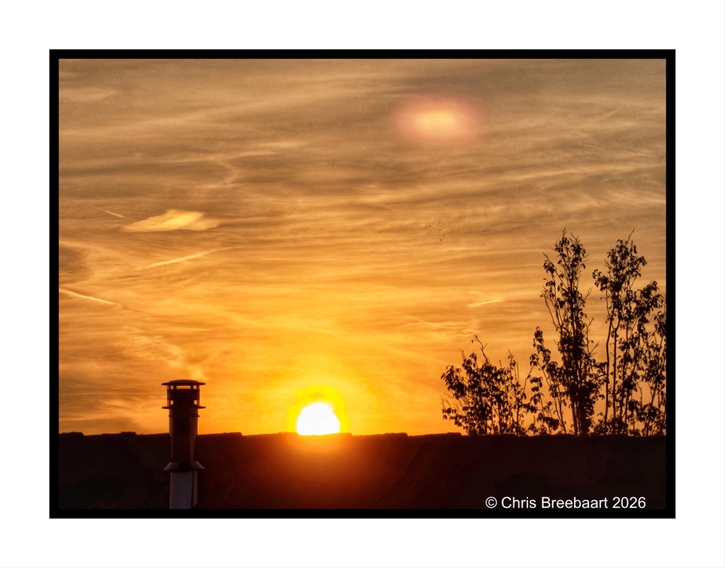 Sunset with vibrant orange and yellow hues, silhouetted trees, and a chimney in the foreground.
