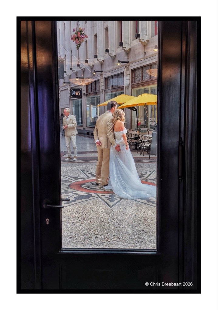 A couple in formal attire stands close together in a courtyard, framed by a doorway. The woman is wearing a flowing white gown, while the man is dressed in a tan suit. In the background, there are people seated at tables under yellow umbrellas, and decorative elements like flowers can be seen.