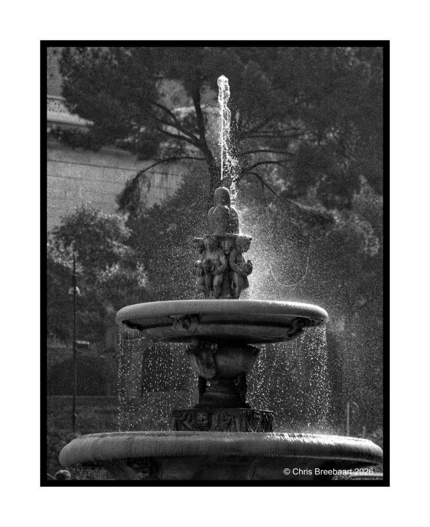 A black and white photograph of a fountain with water spraying from the top, featuring decorative sculptures on the tiered design.