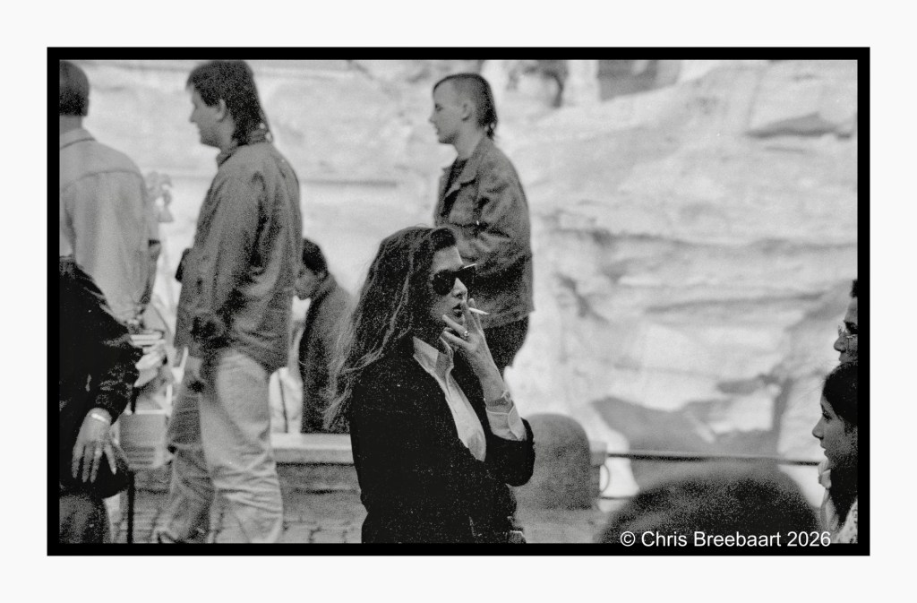 Black and white photograph of a woman in sunglasses smoking a cigarette, surrounded by people in casual clothing. The background features a textured wall.