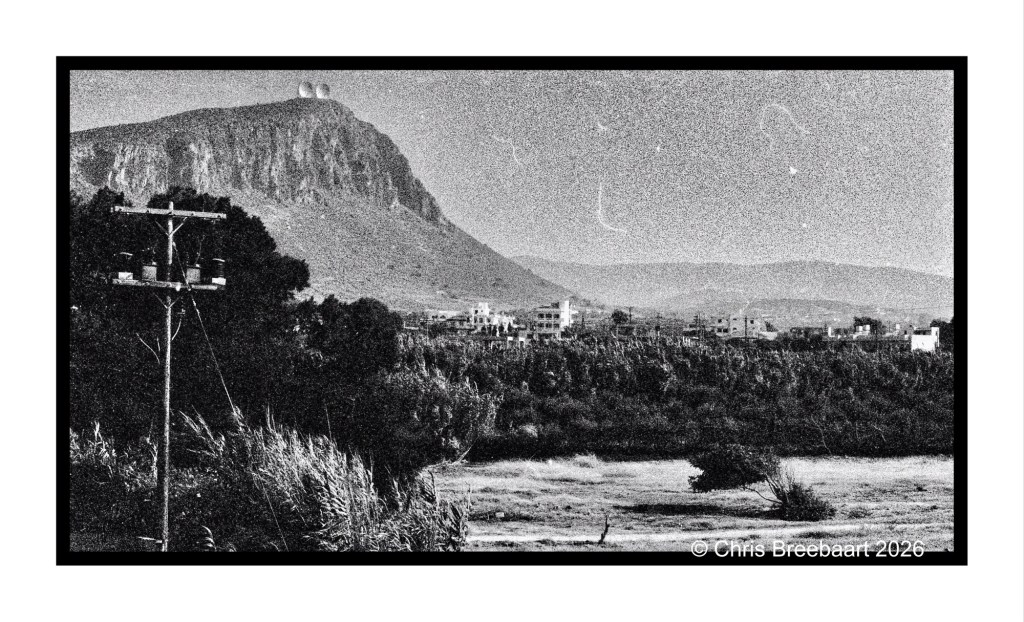 A black and white landscape photograph showing a mountain with two domes at the top, a power pole in the foreground, and a small town in the distance set against a rugged terrain.