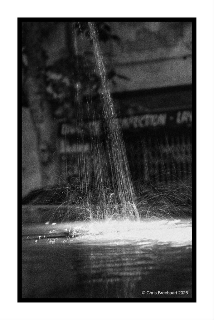A monochrome photo of a fountain with water streaming upwards, creating splashes and ripples on the surface of the pool below.