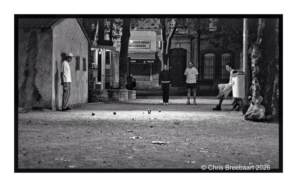 A black and white photograph of a park scene at dusk, featuring four people playing a game, possibly pétanque. One person stands near a building while three others are positioned in the background, engaged in the game. The ground is scattered with leaves, and trees are visible surrounding the area.