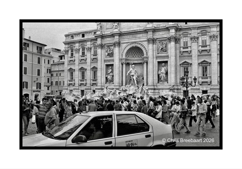 A busy street scene in front of the Trevi Fountain in Rome, featuring a crowd of tourists and a white taxi in the foreground. The image is in black and white.