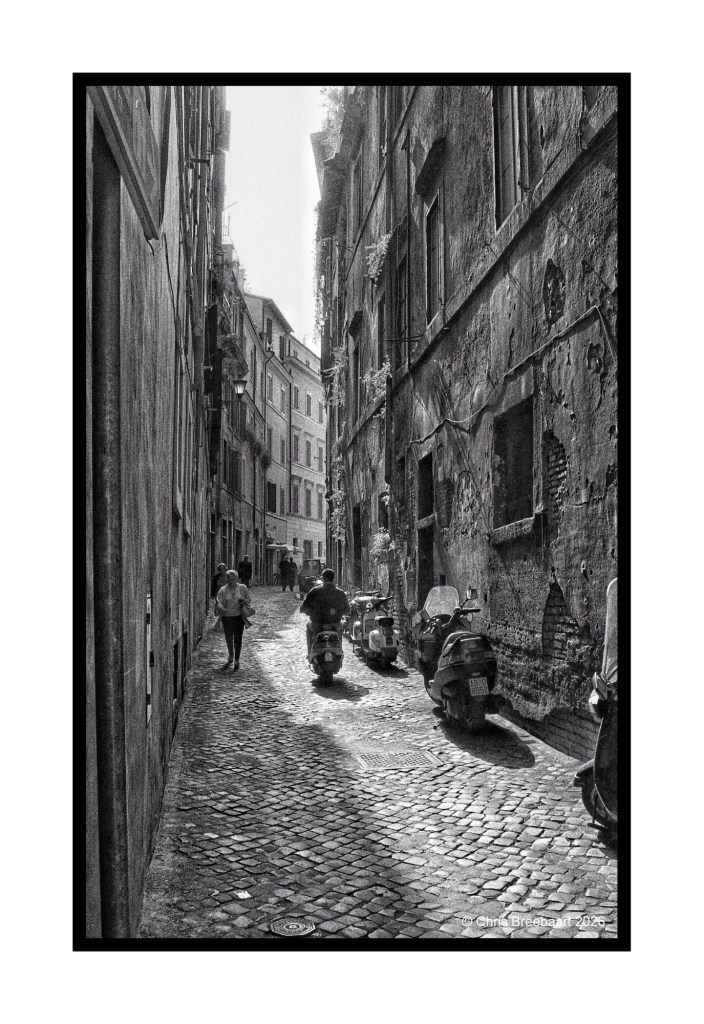 Narrow cobblestone street in a historical city, with weathered buildings on either side and two scooters parked along the curb. A person walks down the street, illuminated by sunlight.