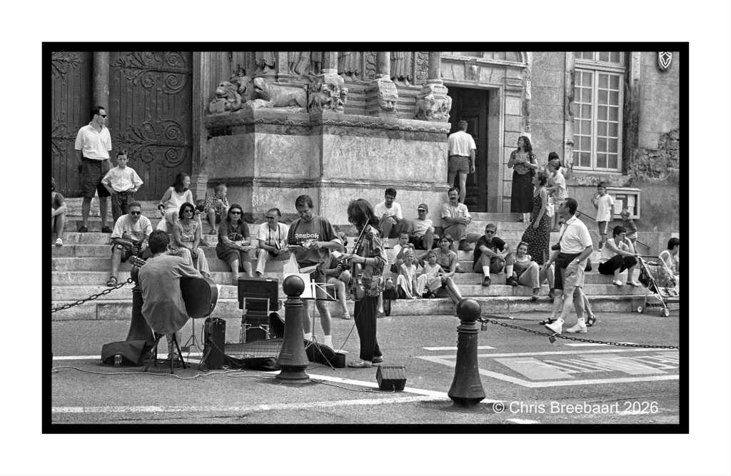 A street musician performing in front of a historic building, surrounded by a diverse audience seated on the steps and nearby sidewalk, creating a lively urban atmosphere.