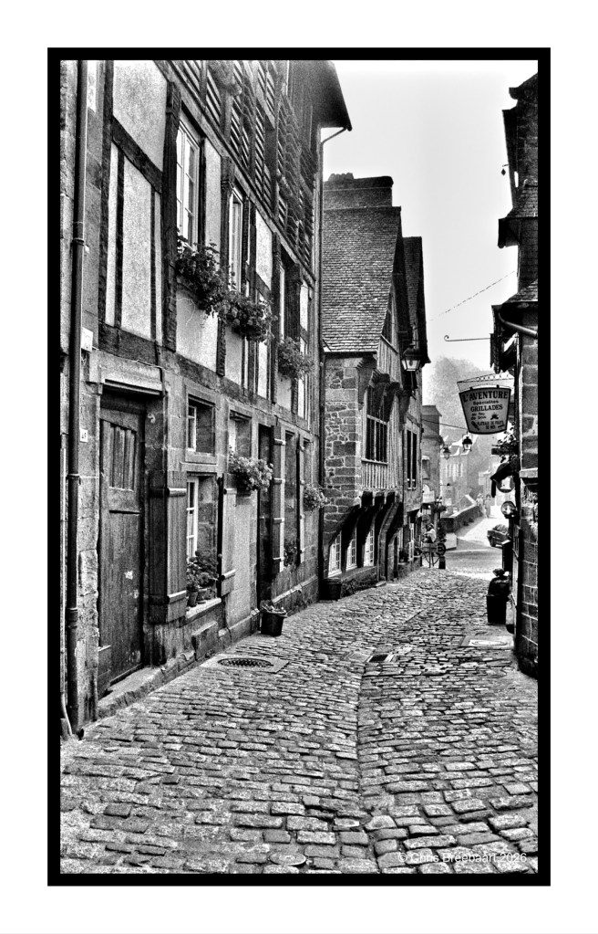 A narrow cobblestone street lined with historic timber-framed buildings, featuring flower boxes. The scene is in black and white, giving it a vintage look.