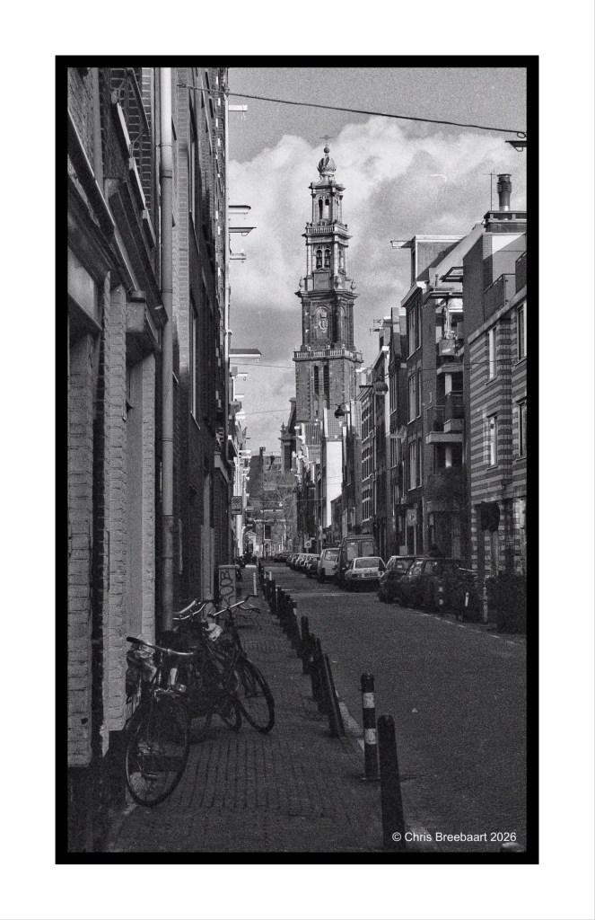A narrow street in a city with bicycles parked on the side, leading towards a tall clock tower in the distance under a cloudy sky.