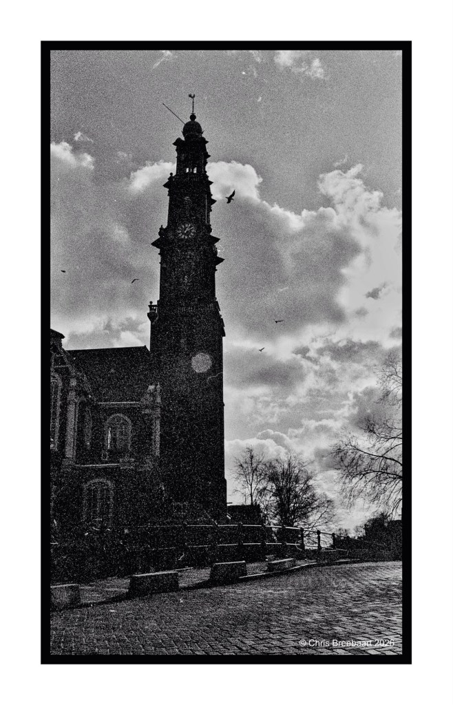 A black and white photograph of a tall clock tower silhouetted against a cloudy sky, with trees and a cobblestone path in the foreground.