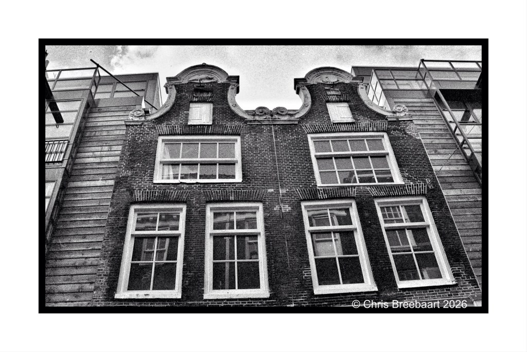 Black and white photograph of an old brick building with large windows and a decorative gable, juxtaposed against a modern glass building.